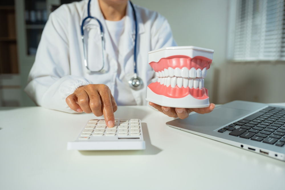 Dentist in a white coat holds a dental tooth model while pointing to a calculator beside a laptop on the desk.