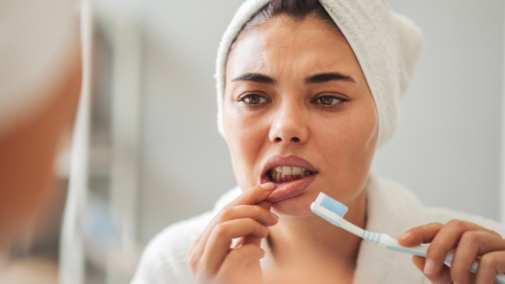 Woman with a towel wrapped around her head brushing her teeth in front of a mirror in a bathroom