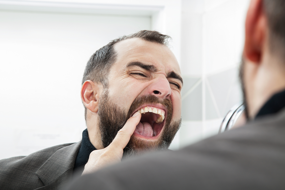Bearded man in a clinical setting opening his mouth wide as another person examines his teeth with a finger in his mouth.