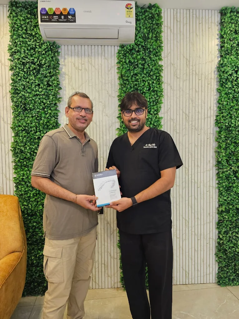 Two men stand indoors as one presents a boxed product to the other, green plant walls in the background.