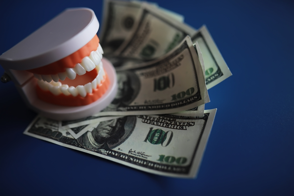 Dental model with an open jaw beside scattered dollar bills on a blue background, implying dental costs or value of care.