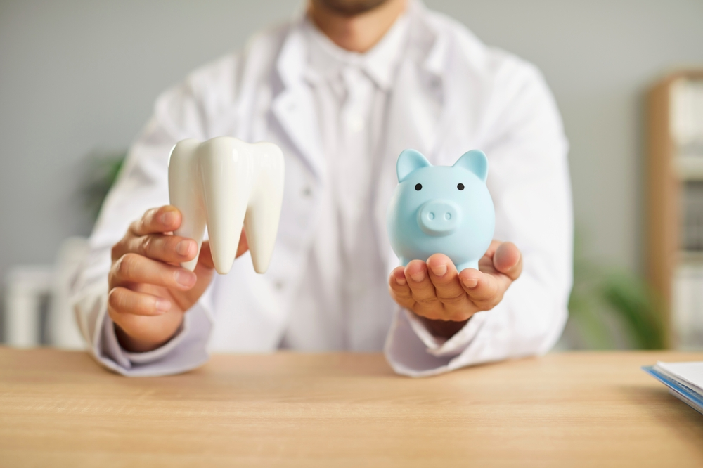 Dentist in a white coat holds a tooth model and a blue piggy bank on a wooden table, illustrating dental care and savings.