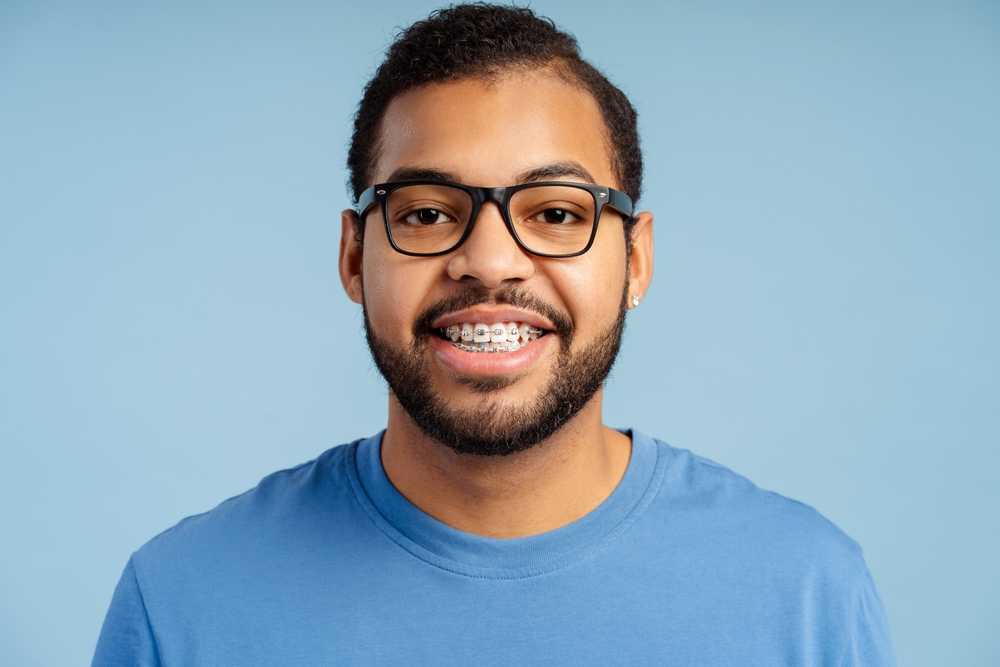Young man with braces and glasses smiling at camera against a blue background.